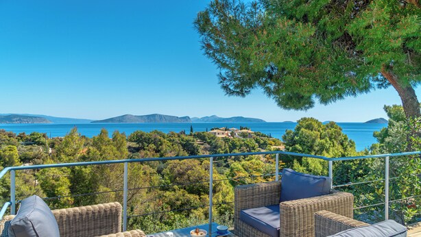 Un coin salon extérieur en osier, avec une table, deux bols et deux verres de jus d’orange, offrant une vue panoramique sur la mer et les collines.
