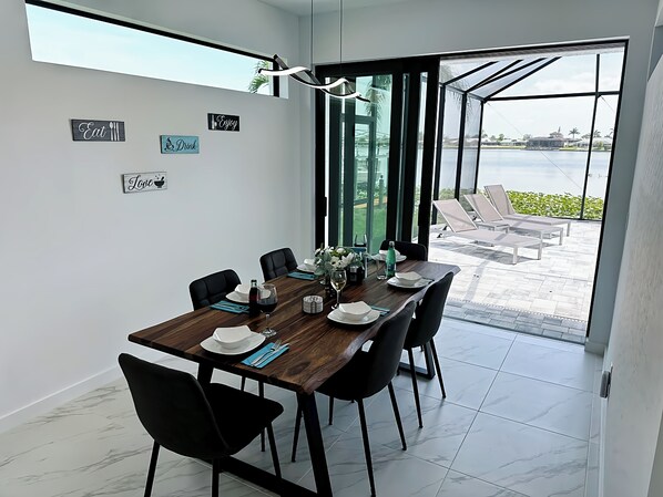 A dining area with a wooden table set for six, black chairs, and a view of a pool and lounge chairs outside. - NEW Exclusive Villa Alhambra Lake - Infinity pool - 5TVs - Gameroom - dock (Cape Coral)
