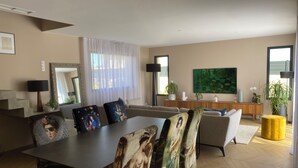 A modern living room with a dining area, featuring a large flat-screen TV, a sofa set, a coffee table, and a dining table with chairs. - Maison / Piscine, Proche Plages (Vic-la-Gardiole)