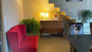 A living room with a red sofa, a wooden cabinet, a dining table, and a staircase with a painting above it. - Maison / Piscine, Proche Plages (Vic-la-Gardiole)