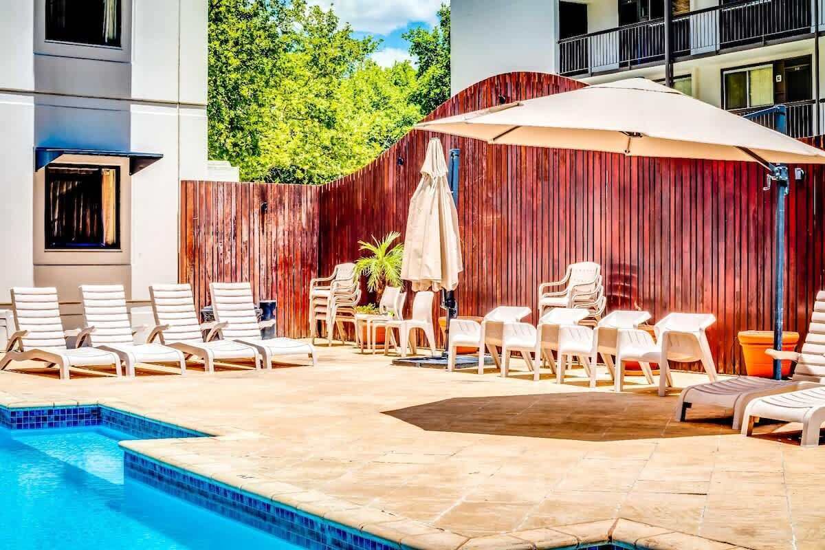 A pool area with lounge chairs and umbrellas, surrounded by a wooden fence and a building with balconies.