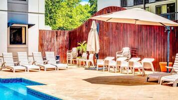 A pool area with lounge chairs and umbrellas, surrounded by a wooden fence and a building with balconies.