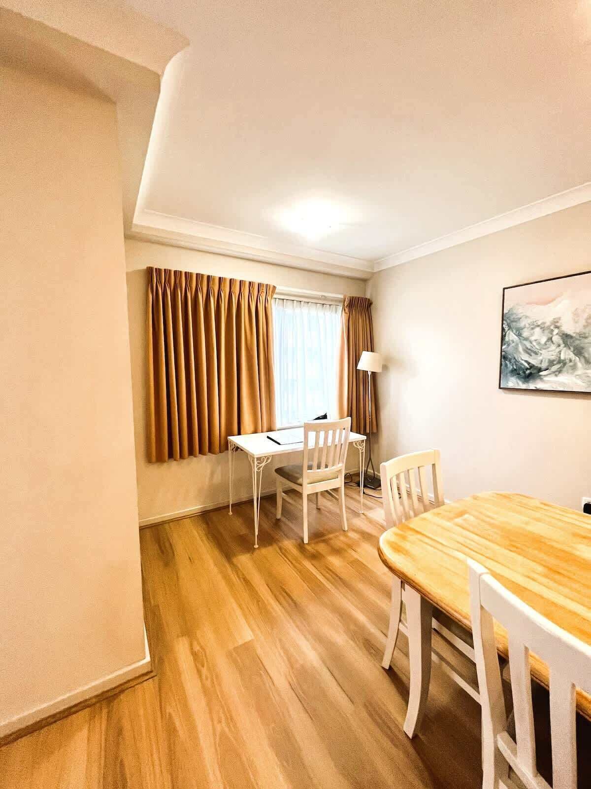 A room with a wooden dining table, white chairs, a white desk, and a window with brown curtains.