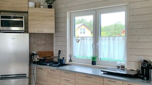 Moderni keittiö, jossa on ruostumattomasta teräksestä valmistettu jääkaappi, puukaapit, sisäänrakennettu uuni ja ikkuna, josta avautuu näkymä taloon ja vehreään maisemaan.