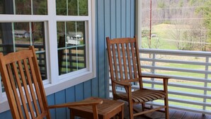 En träveranda med två stolar och ett litet bord, med utsikt över en inhägnad trädgård med träd och ett hus i fjärran.