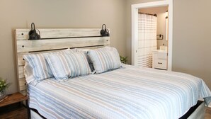 Un dormitorio con cabecera de madera, ropa de cama a rayas, lámparas de noche, una mesita de noche y vistas al baño.