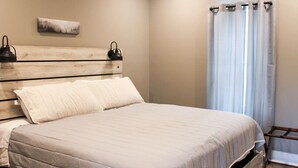 A bedroom with a wooden headboard, a bed with white bedding, a bedside table with a lamp, and a window with curtains. - Tiny 2br Cabin w/ View | Grill | Fire Pit #3 (West Jefferson)