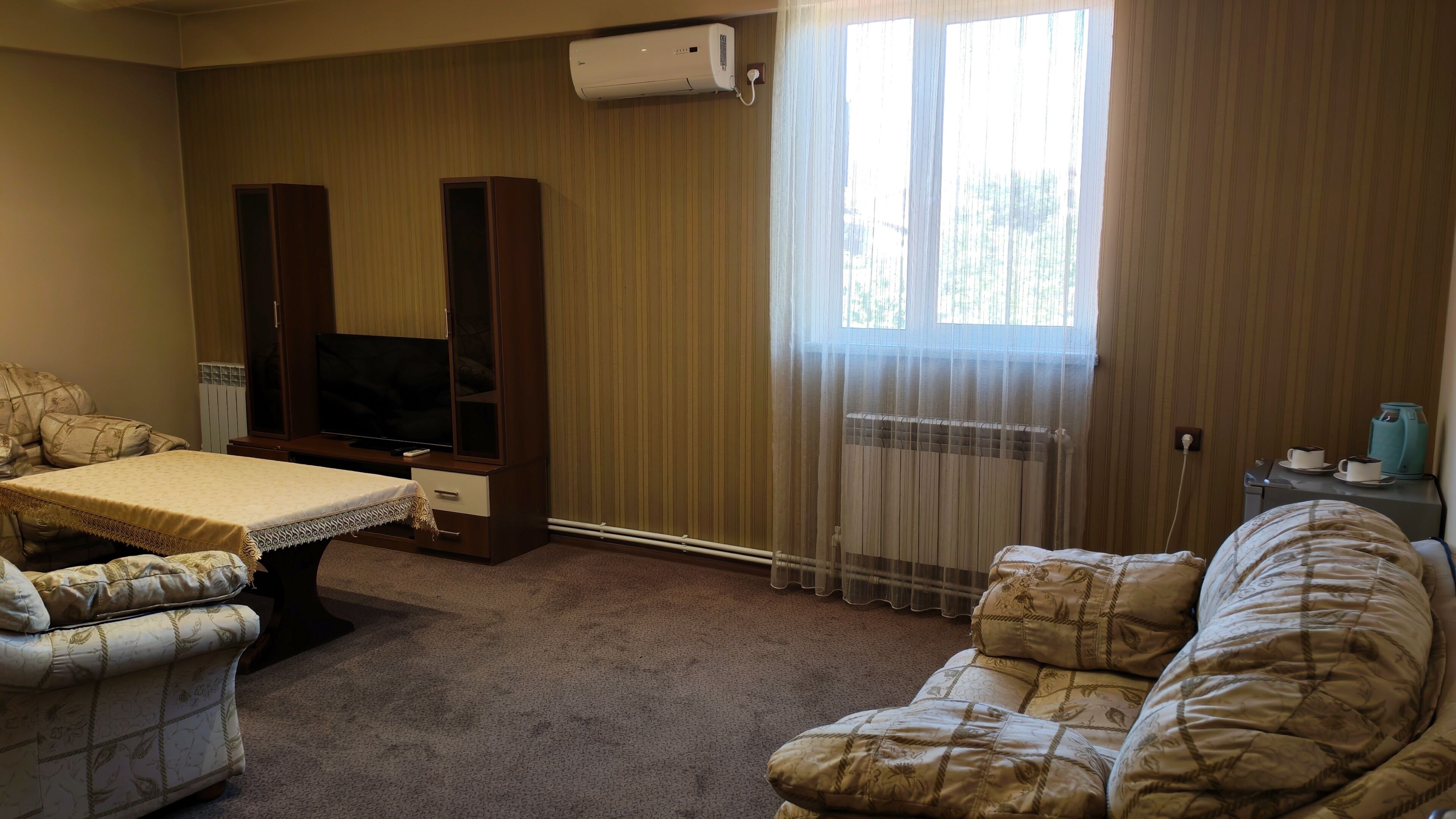 A hotel room with a bed, a television, a sofa, a small table with a kettle, and a window with curtains.