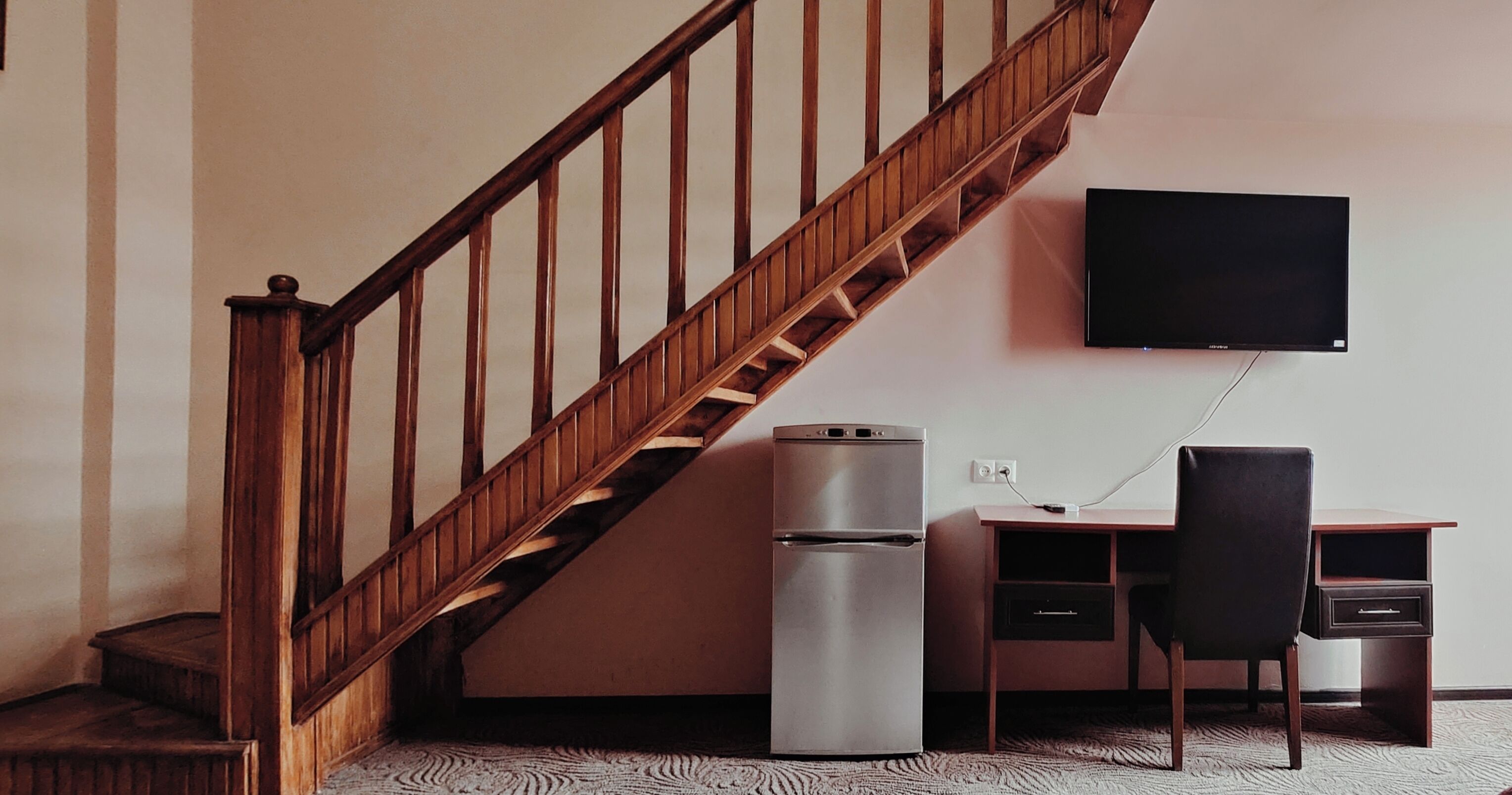 Een kamer met een houten trap, een koelkast, een bureau met stoel, en een wandgemonteerde flatscreen televisie.