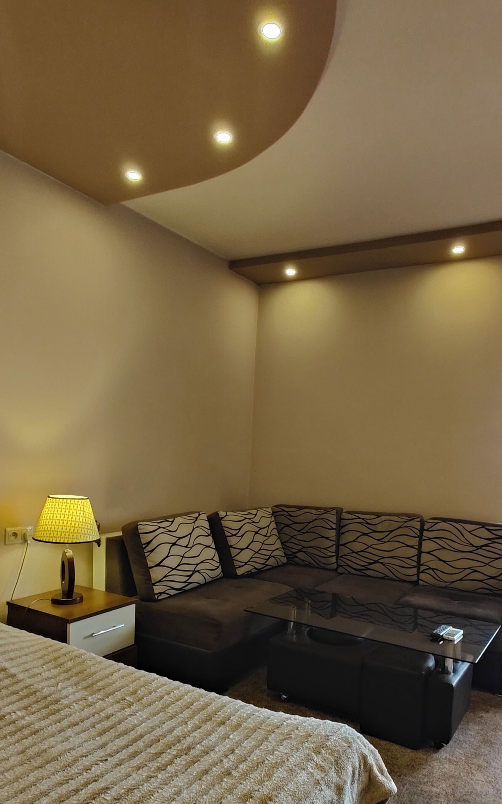 A modern hotel room with a sectional sofa, a glass coffee table, a lamp, and a bed with a striped blanket.
