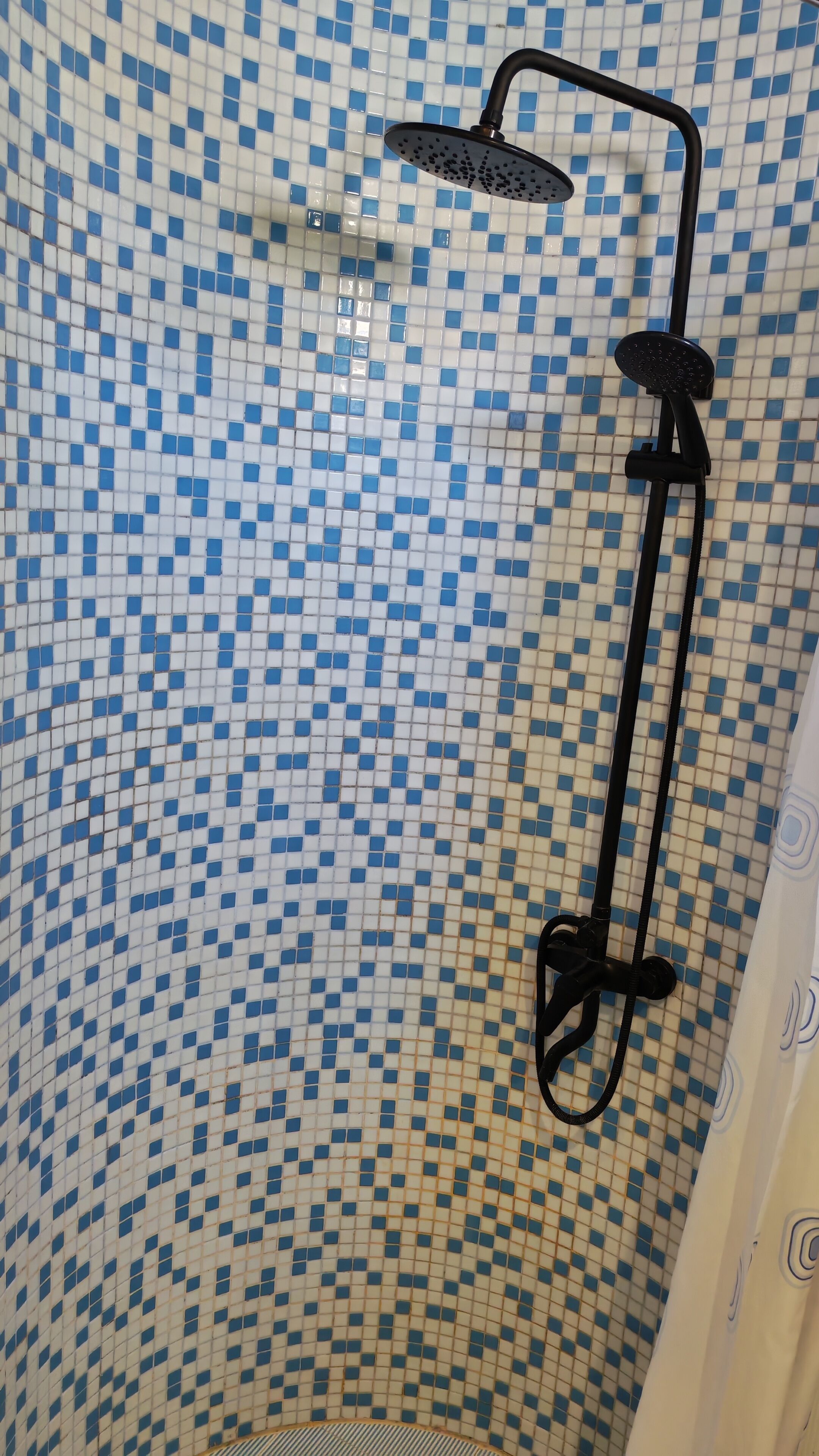 Ducha con manguera negra y cabezal de ducha manual, frente a una pared de azulejos con un diseño azul y blanco.