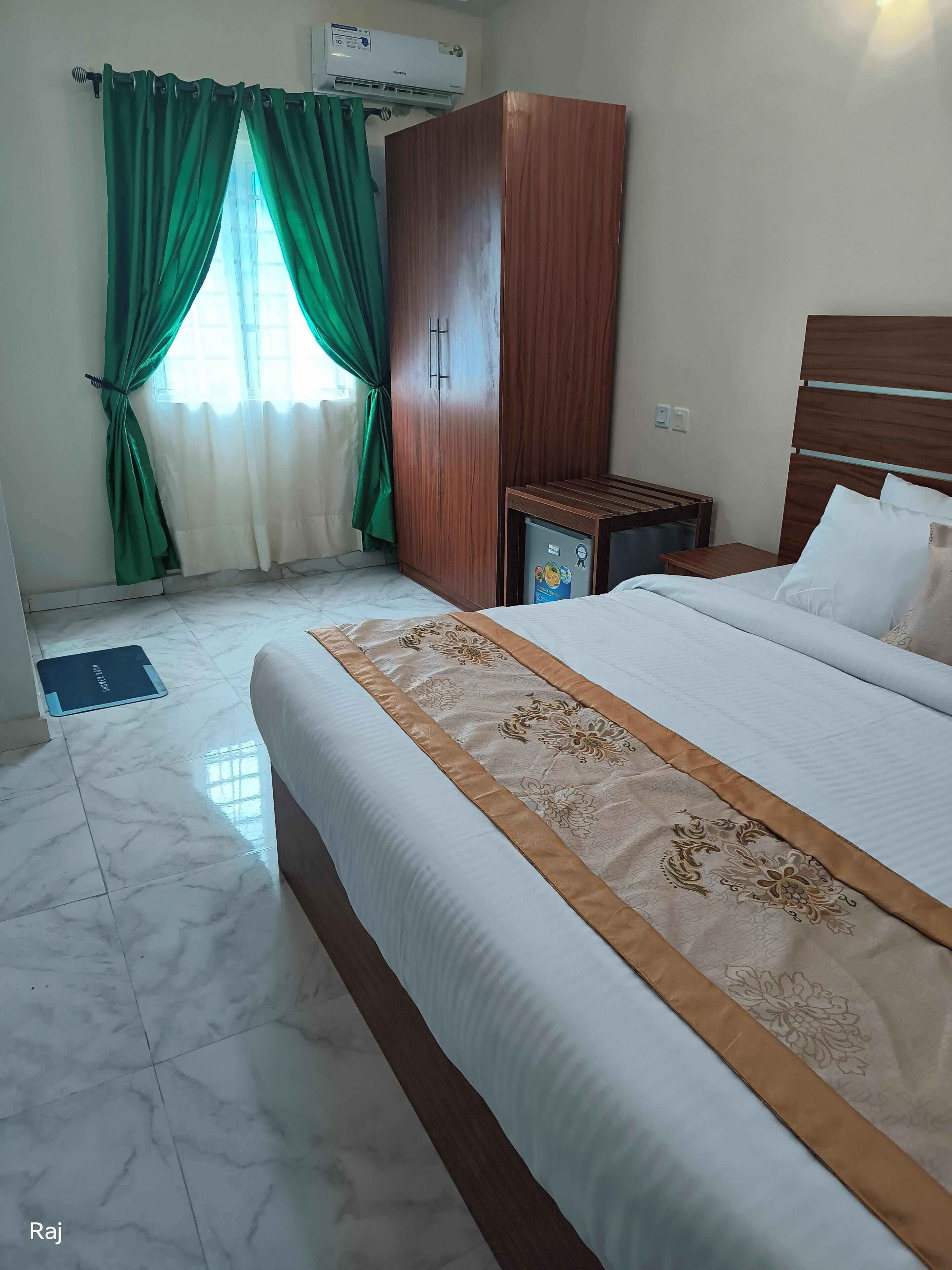 Una habitación de hotel con cama, armario, mesita de noche y ventana con cortinas verdes.