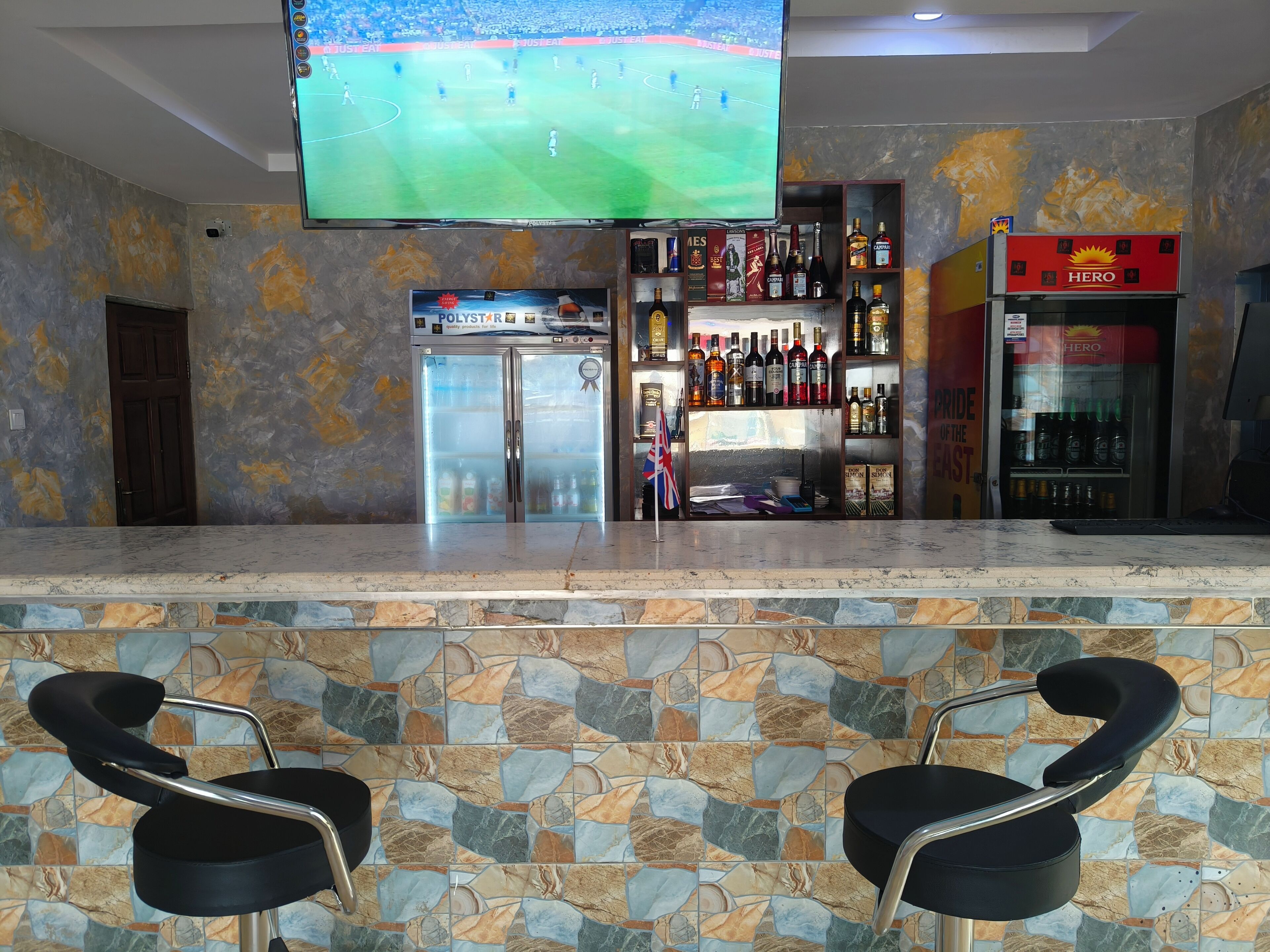 A bar area with a television displaying a sports event, a refrigerator, shelves with bottles, and two bar stools.