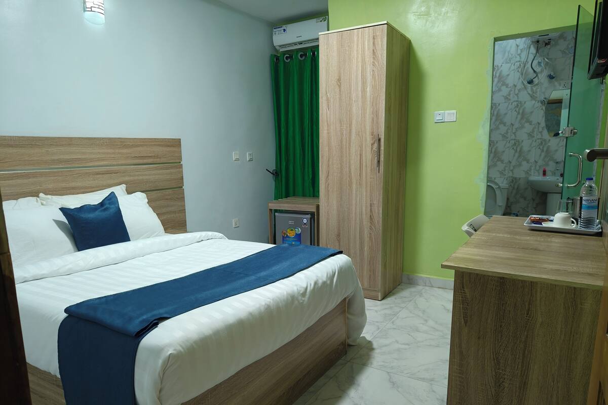 A neatly arranged hotel room with a bed, a wardrobe, a small table with a water bottle, and a green curtain.