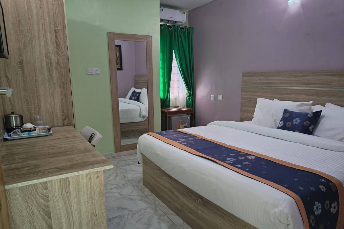 A hotel room with a wooden desk, a bed with a floral bedspread, a nightstand, and a green curtain.