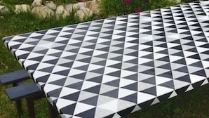 Una mesa de picnic con mantel a cuadros blancos y negros, rodeada de follaje y un edificio al fondo.