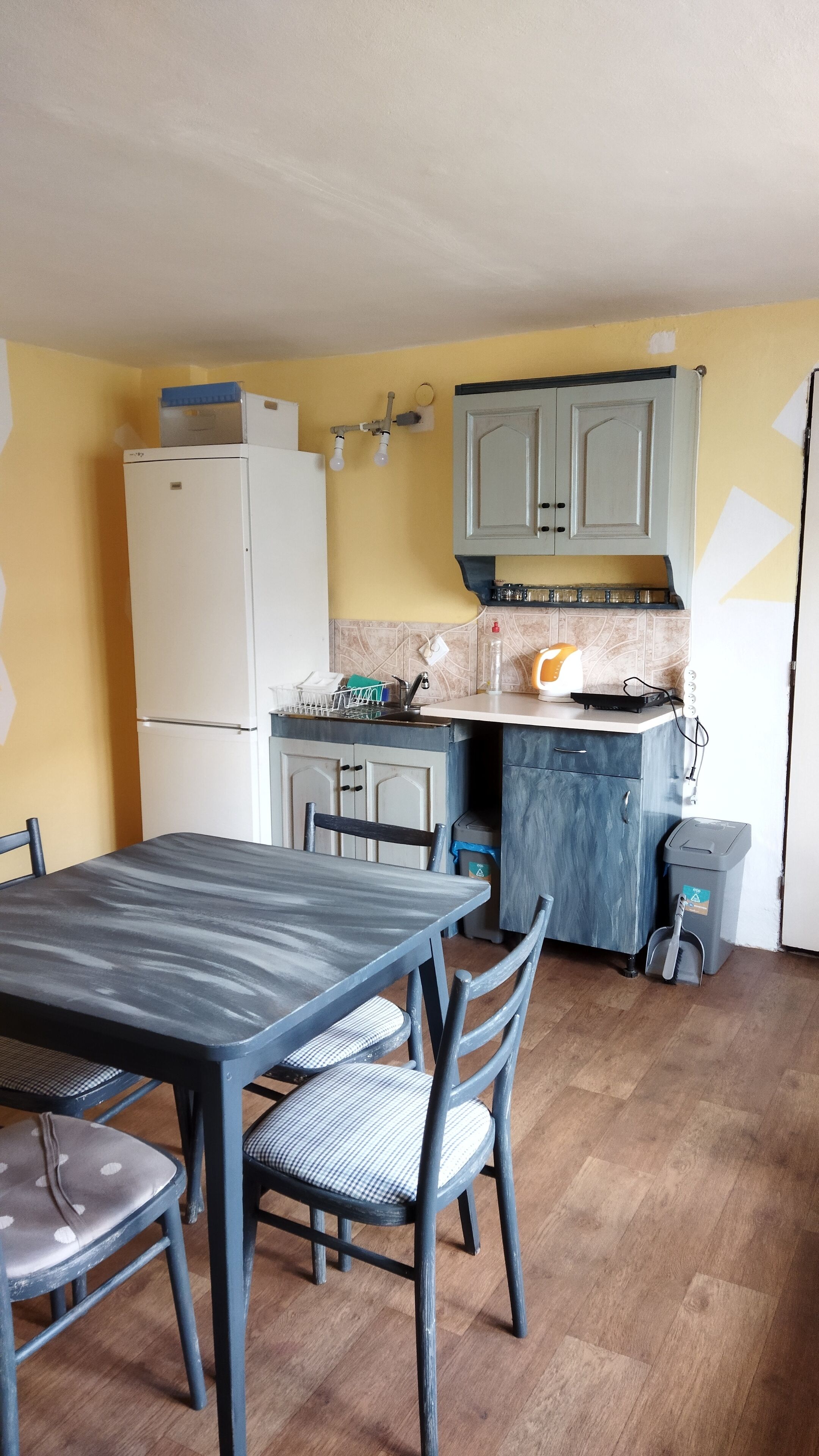 Eine kleine Küche mit Kühlschrank, Spüle und einer Arbeitsplatte mit Wasserkocher. Ein Esstisch mit Stühlen und einer Mülltonne sind ebenfalls vorhanden.