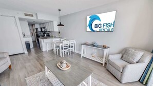 Et moderne køkken med hvide skabe, et spiseområde med bord og stole, og en opholdsstue med sofa og tv, der viser 'BIG FISH RENTALS'.