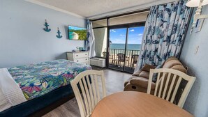 Quarto com uma cama, uma mesa de madeira e um sofá. Há uma varanda com vista para o mar.