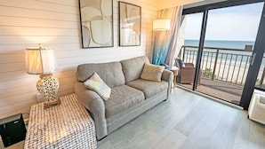 A modern living room with a grey sofa, patterned ottoman, abstract wall art, and a balcony with a view of the ocean. - New Remodeled, Grand Cayman, Amazing Decor, Pools! (Myrtle Beach)