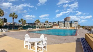 Un espace piscine avec des chaises blanches et une table, entouré de palmiers et d’un bâtiment doté de nombreux balcons.