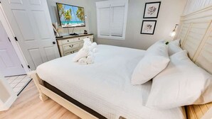 Una cama bien hecha con sábanas y almohadas blancas, un televisor en una cómoda y una ventana con persianas.