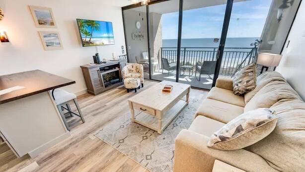 Una sala moderna con sofá, mesa de centro y televisor. Tiene un balcón con vista al mar.