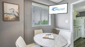 A room with a round table, white chairs, a framed picture, a window with blinds, and a TV screen displaying 'BIG FISH RENTALS'.