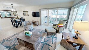 A spacious living room with a large window overlooking a beach, a dining area with a round table and chairs, and a cozy seating area with a lamp.