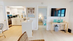 A modern kitchen with white cabinetry, a central island, and a dining area with wicker furniture. A television and decorative items are visible in the living space. - Crescent Arms Vacation Rentals 203N Siesta Key Florida (Sarasota)