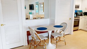 A dining area with a white table and two wicker chairs, a kitchen with white cabinets and a microwave, and a door with a golden handle. - Crescent Arms Vacation Rentals 203N Siesta Key Florida (Sarasota)