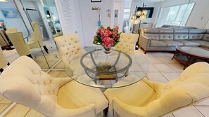 Uma sala de estar moderna com uma mesa de centro de vidro, rodeada por poltronas bege. Um vaso com flores cor-de-rosa está sobre a mesa.