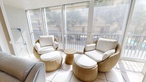 A sunlit balcony with wicker furniture and a view of a pool and buildings.