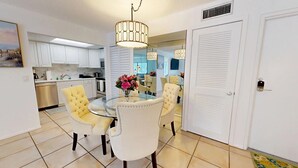A modern kitchen with white cabinets, a stainless steel dishwasher, and a dining area with a glass table and yellow chairs.