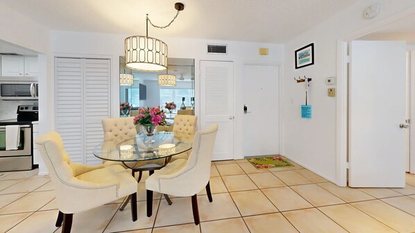 A dining area with a glass table and four chairs, a chandelier, and a kitchen in the background.