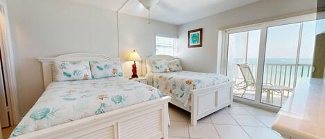 A room with two beds, a ceiling fan, a window with blinds, and a view of the ocean through a balcony.