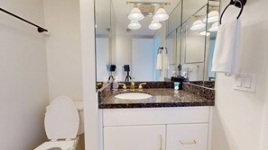 Un baño con inodoro blanco, lavabo de encimera de granito, grifería dorada y un espejo grande.
