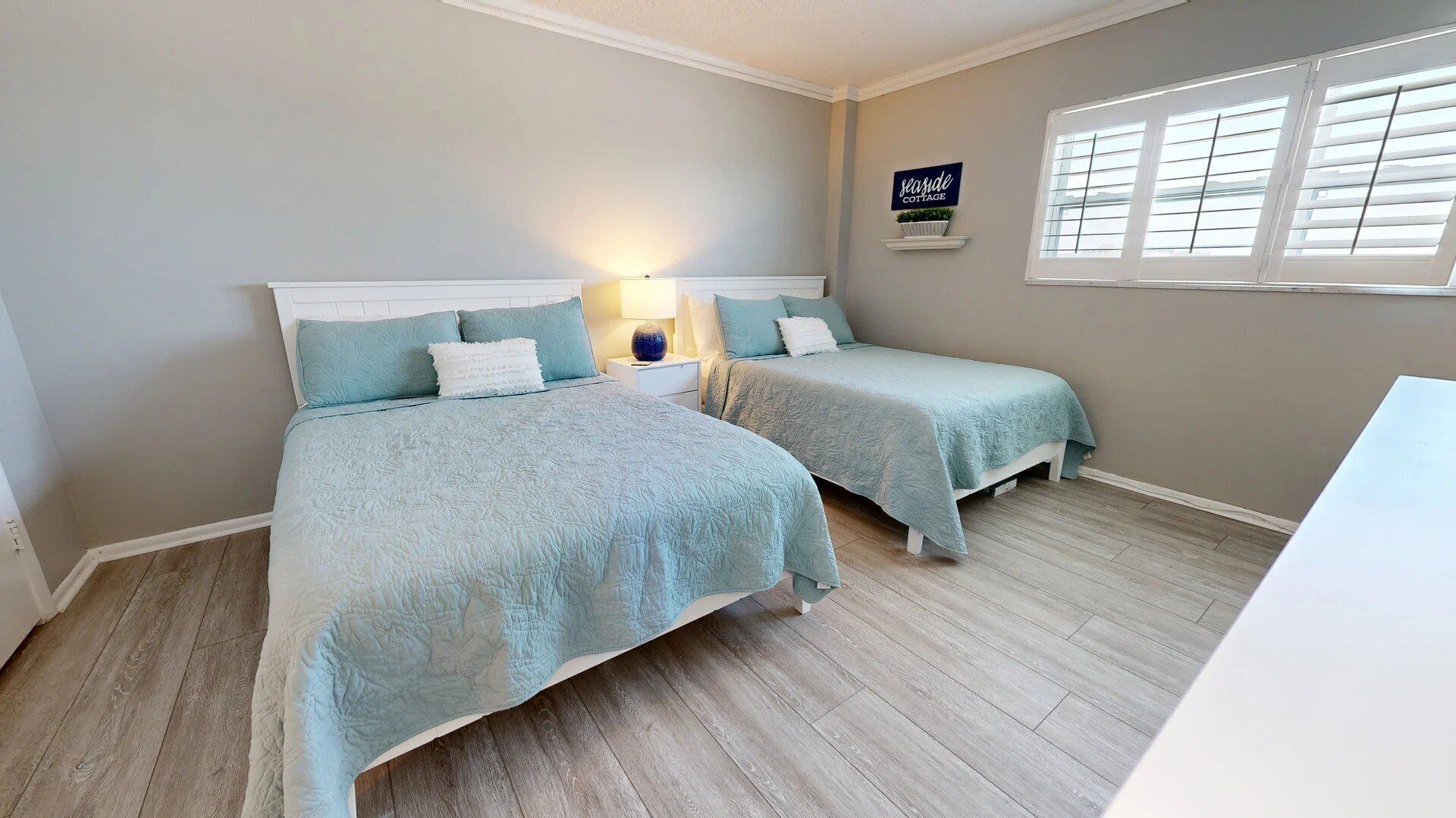 Quarto com duas camas, mesa de cabeceira, abajur e janela com persianas.