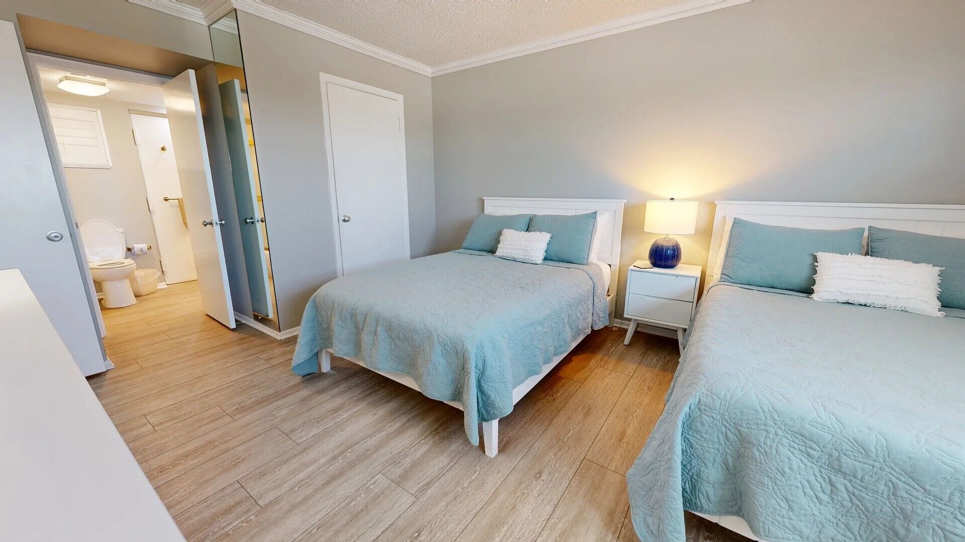 Habitación de hotel con dos camas, una mesita de noche con lámpara, y un baño visible a través de una puerta abierta.