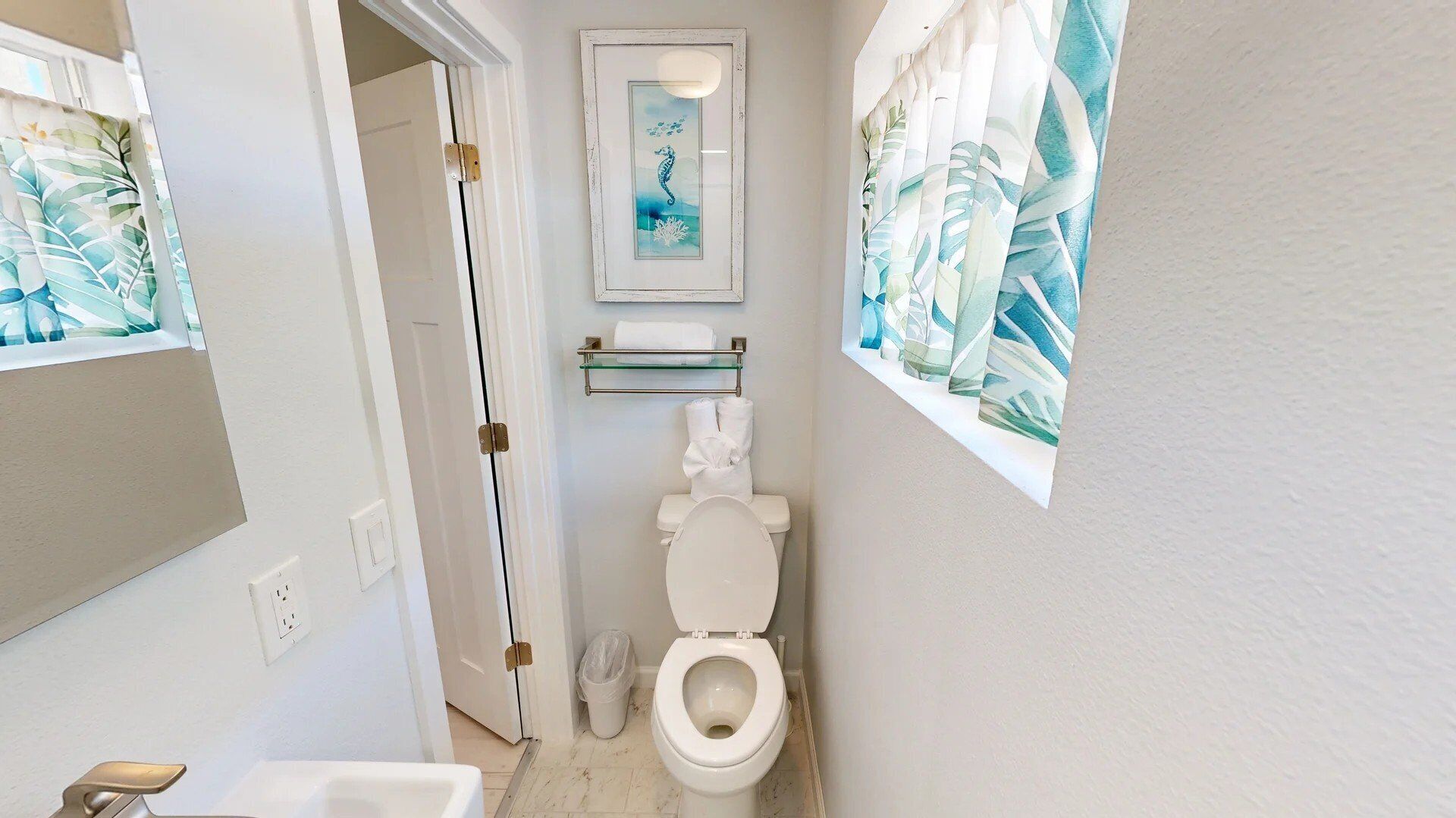 A small bathroom with a toilet, a glass shelf above the toilet, and a framed seahorse artwork on the wall.