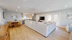 A modern living room with a large L-shaped sofa, a dining area with a wooden table and chairs, a kitchen with white cabinets, and a ceiling fan.