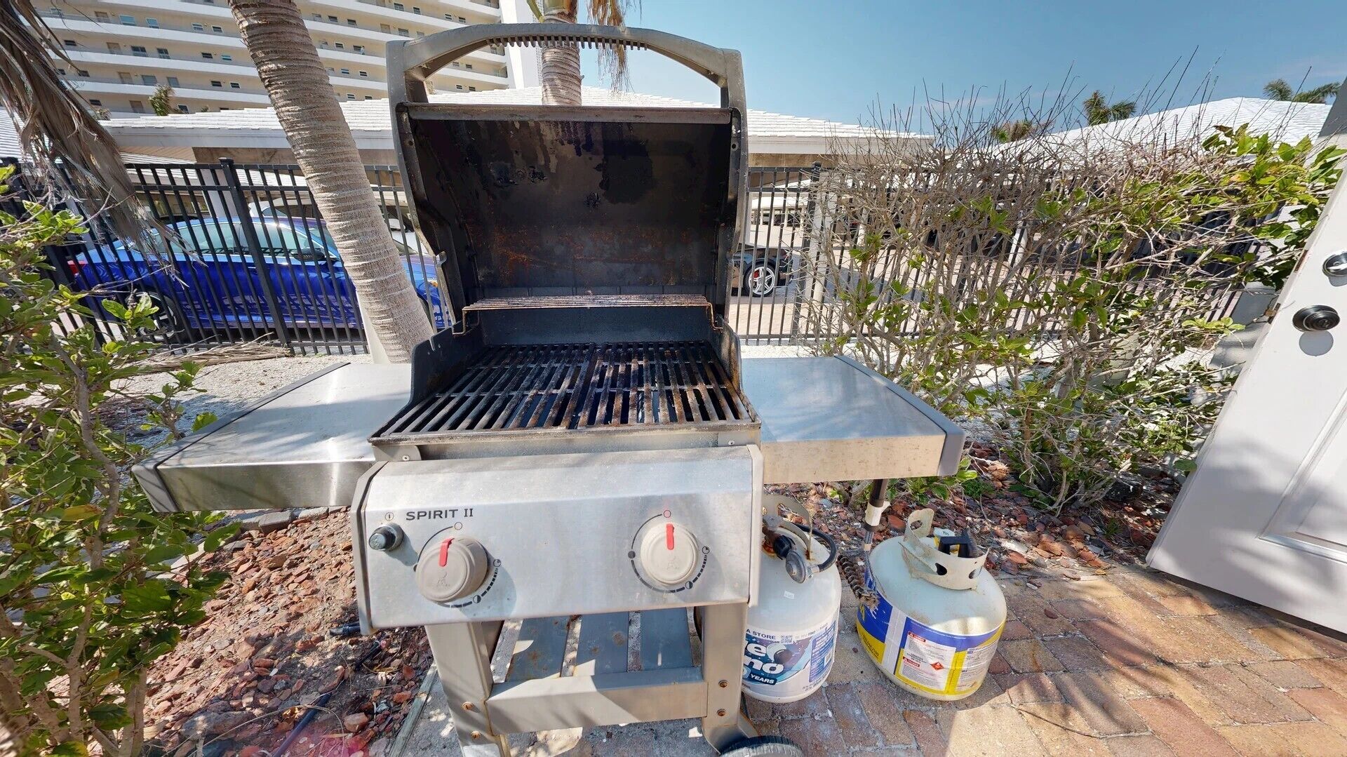 Un barbecue au gaz portatif, équipé de deux bouteilles de propane, installé à l’extérieur, avec un bâtiment et des palmiers en arrière-plan.