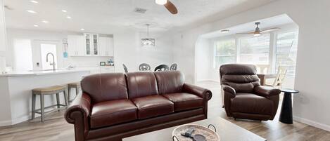 A spacious living room with a brown leather sofa, a matching armchair, and a coffee table with a decorative tray. A ceiling fan and recessed lighting are visible.