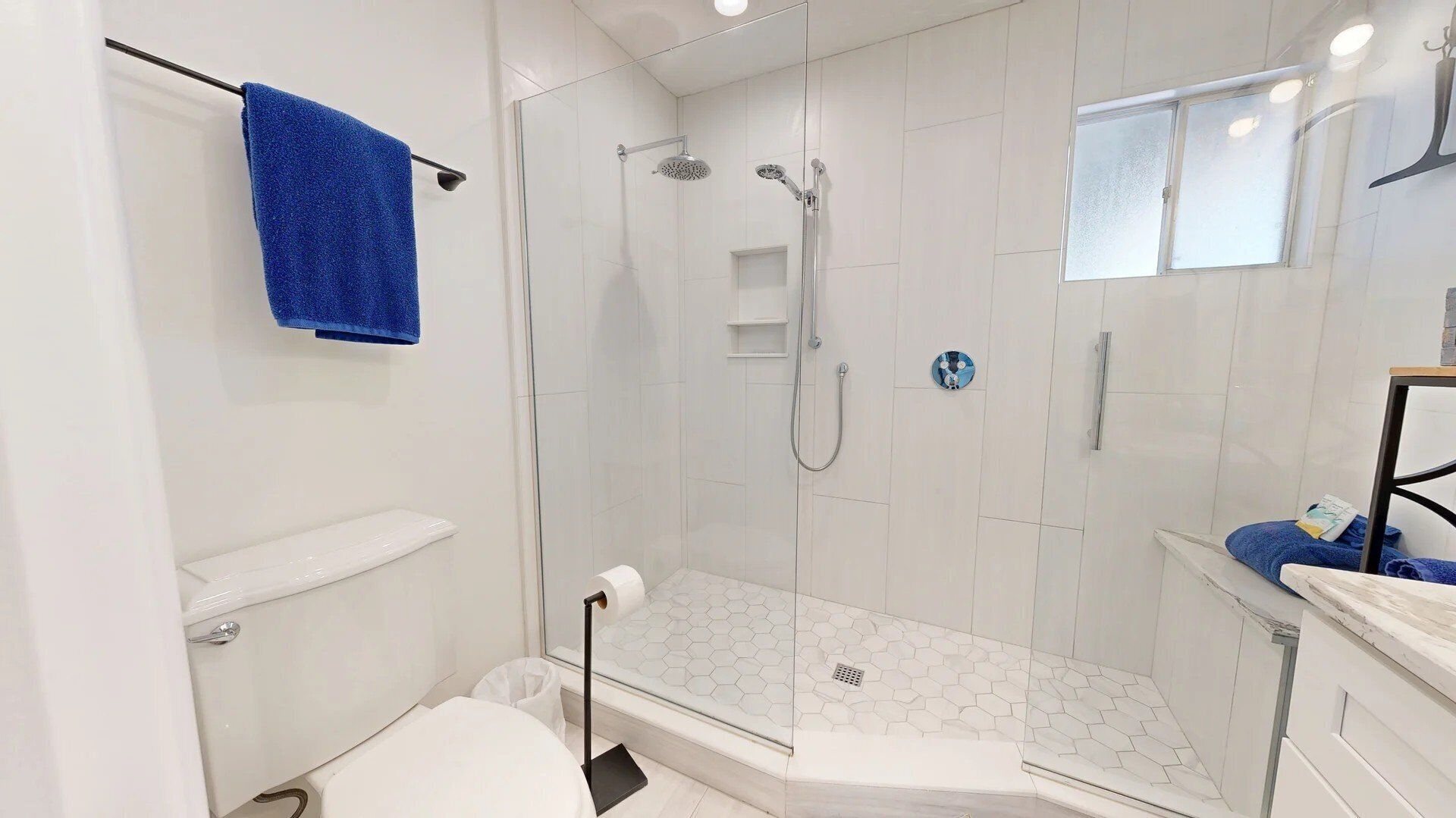 Une salle de bain moderne avec une douche à parois de verre, des carreaux blancs, une serviette bleue et une petite fenêtre.