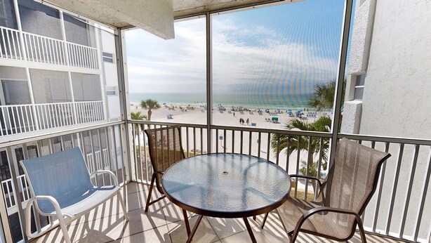 Ein Balkon mit Glastisch und Stühlen, der auf einen Strand mit Sonnenschirmen und Menschen blickt.