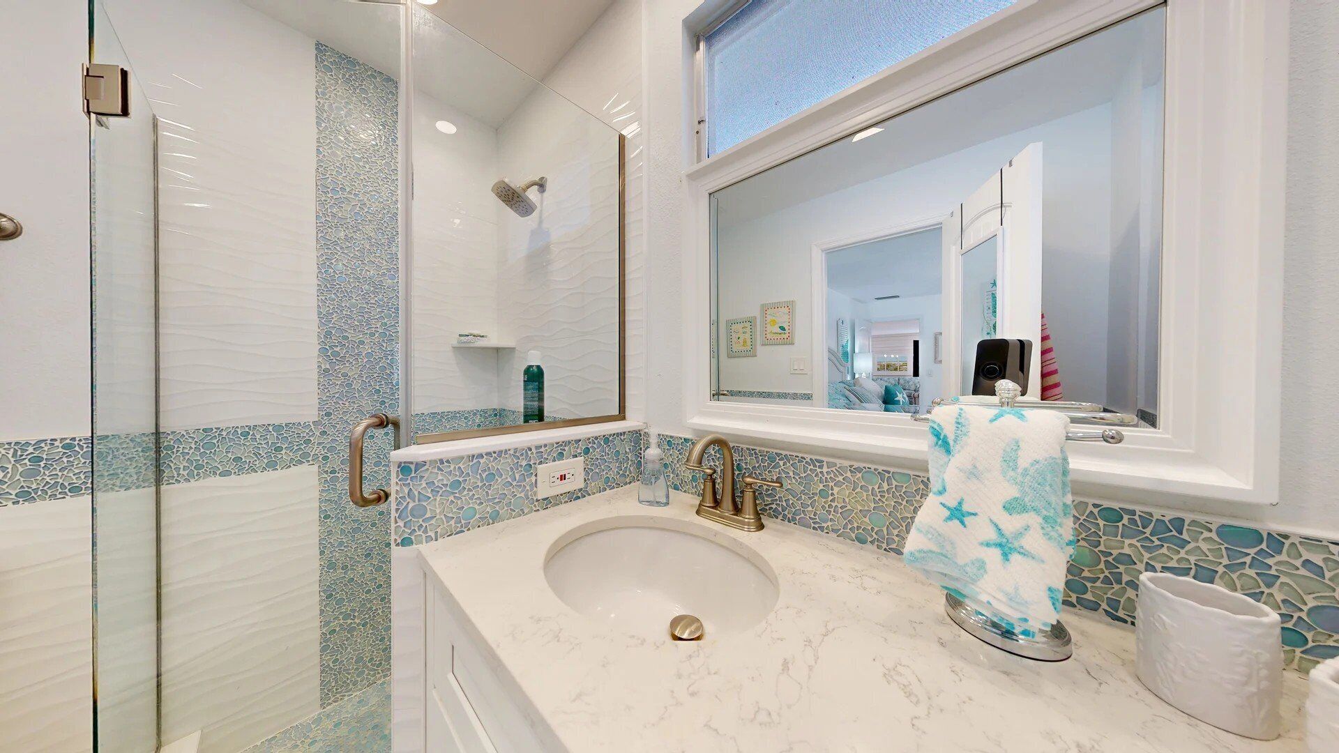 A bathroom with a glass shower enclosure, a sink with a marble countertop, and a mirror with a view of the adjacent living area.