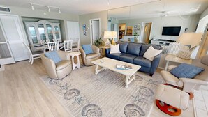 Un salon spacieux avec un canapé bleu, des fauteuils beige et une table basse. On y trouve également un coin repas avec des chaises blanches et un buffet. La pièce est dotée d’un grand miroir et d’un téléviseur.