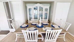 Une salle à manger avec une table en bois dressée pour six personnes, des chaises blanches et, au fond, un buffet blanc aux portes en verre.