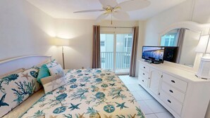 A bedroom with a bed, a TV, a ceiling fan, a balcony, and a window with curtains. - Crescent Arms Vacation Rentals 405S Siesta Key Florida (Sarasota)