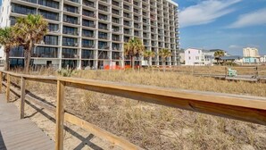 A multi-story hotel with a grid of balconies, a wooden boardwalk in the foreground, and a sandy beach area. - Octopus Garden: 1BR, King Bed, Indoor/Outdoor Pool (Carolina Beach)